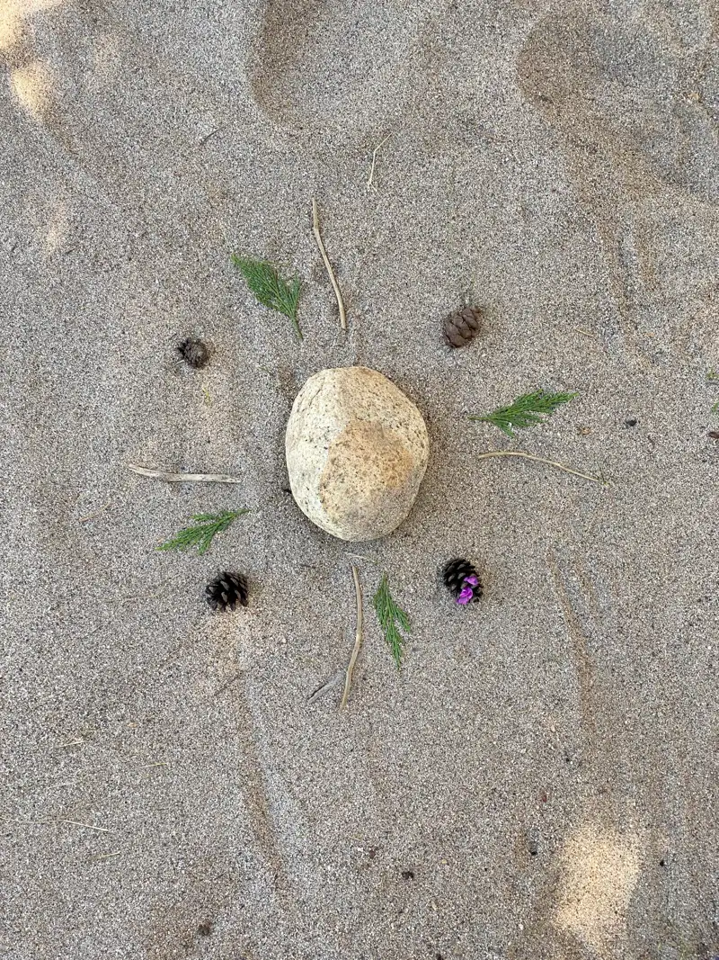 A stone surrounded by small leaves, twigs, and pine cones arranged in sand.